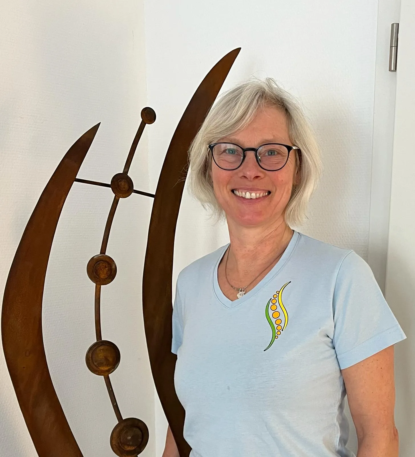Anke Sewald, lächelnd im Praxishemd vor einer geschwungenen Metallskulptur.